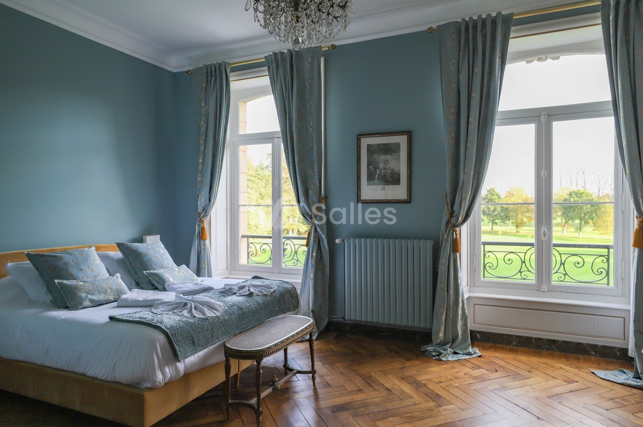 Chambre lumineuse avec lit double, parquet en bois, murs bleus, grandes fenêtres donnant sur un jardin verdoyant.