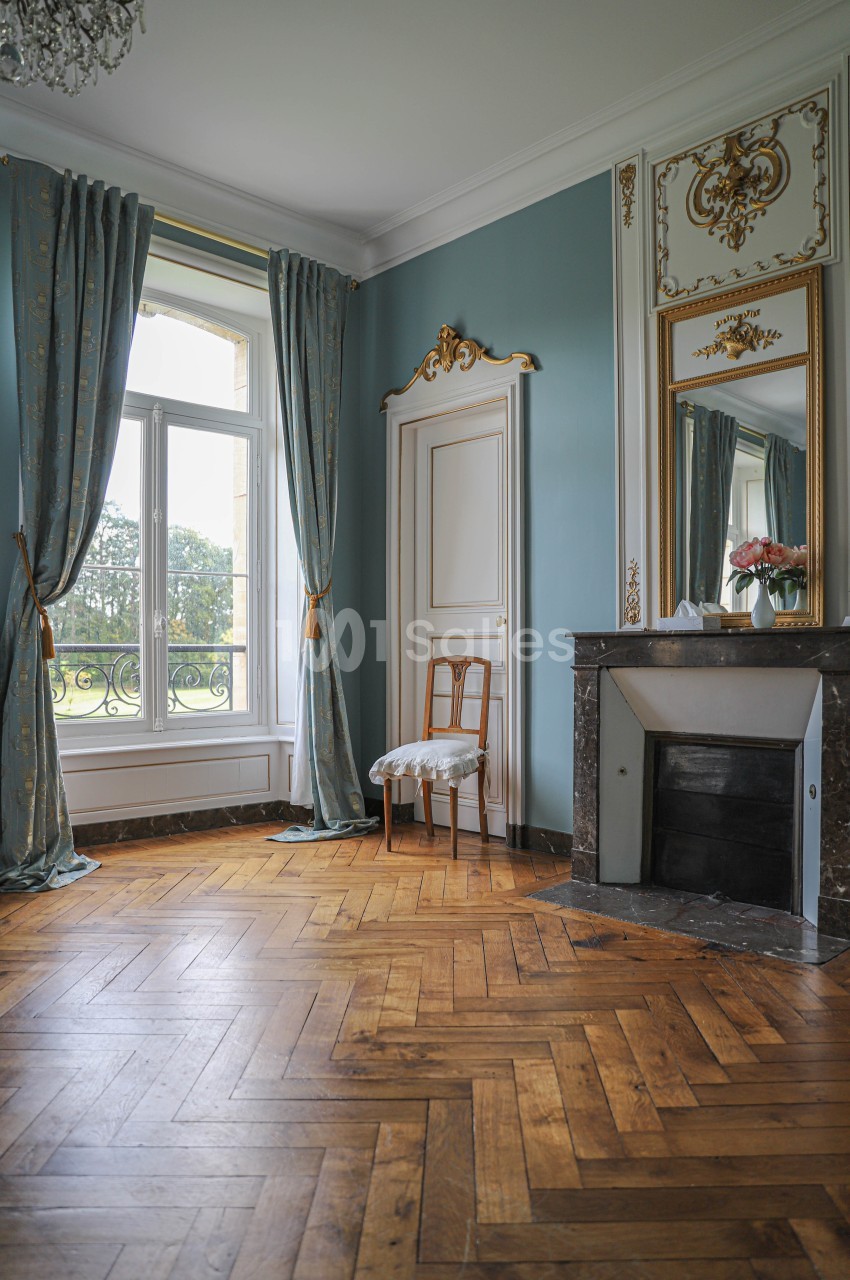 Pièce lumineuse avec parquet en bois, cheminée en marbre, miroir doré, rideaux bleus et vue sur un jardin.