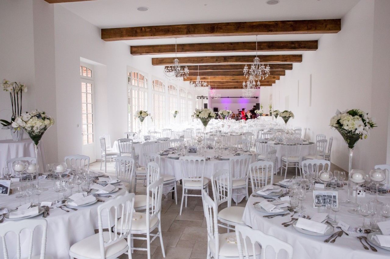 Salle de réception lumineuse avec tables décorées de nappes blanches, chaises assorties et arrangements floraux élégants.