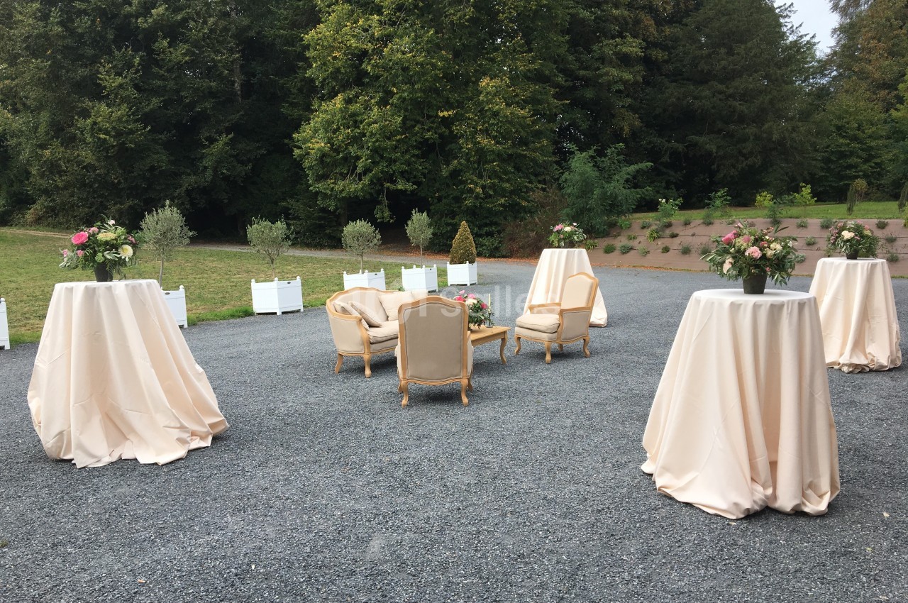 Mobilier de réception avec fauteuils et tables hautes drapées, disposés en extérieur sur un sol gravillonné.