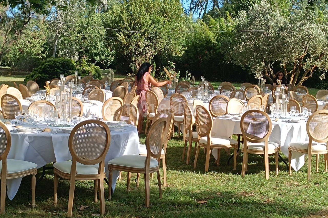 Tables rondes dressées avec nappes blanches et chaises en bois dans un jardin, préparées pour un événement en plein air.