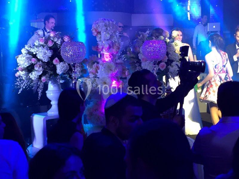 Salle de réception avec invités, grands bouquets de fleurs et éclairage bleu, un photographe au premier plan.