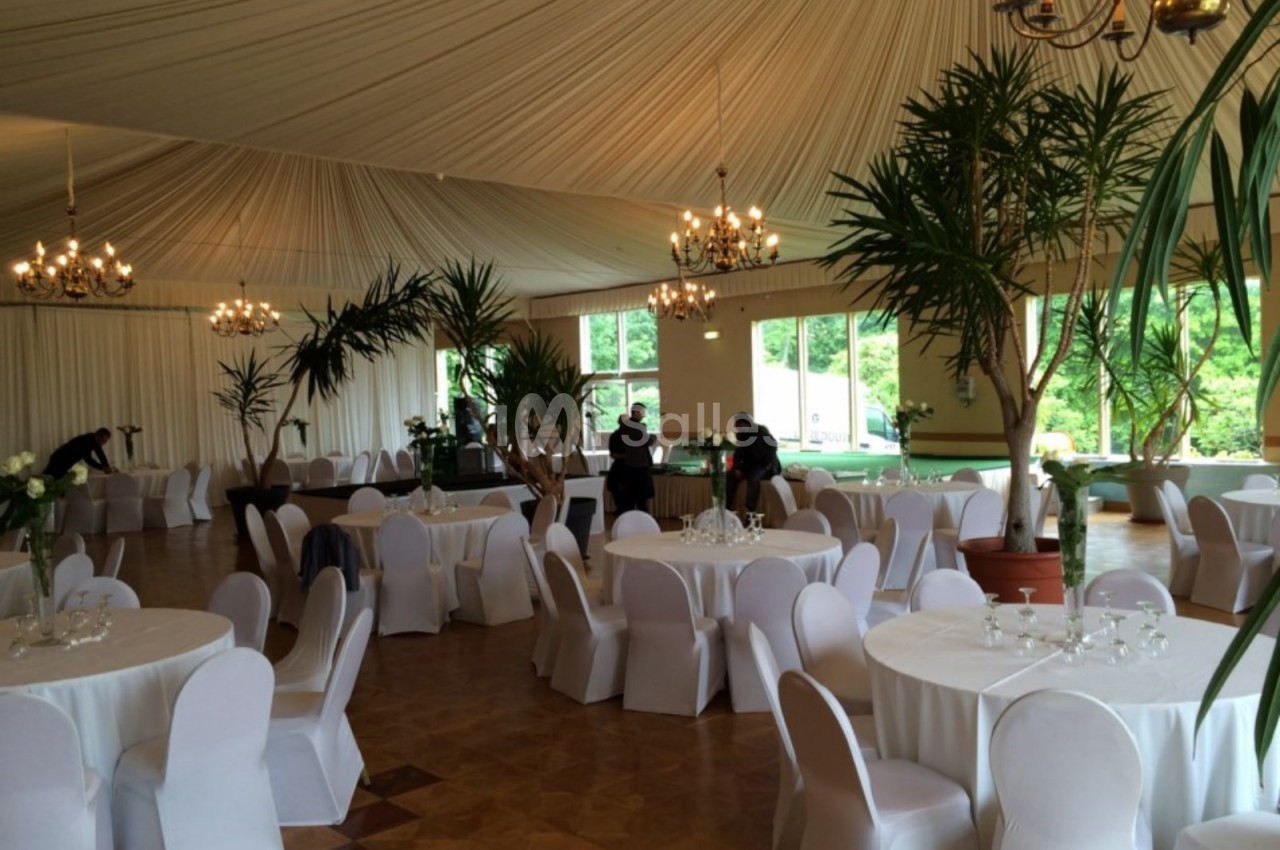 Salle de réception élégante avec tables rondes dressées, chaises blanches et grandes plantes décoratives.