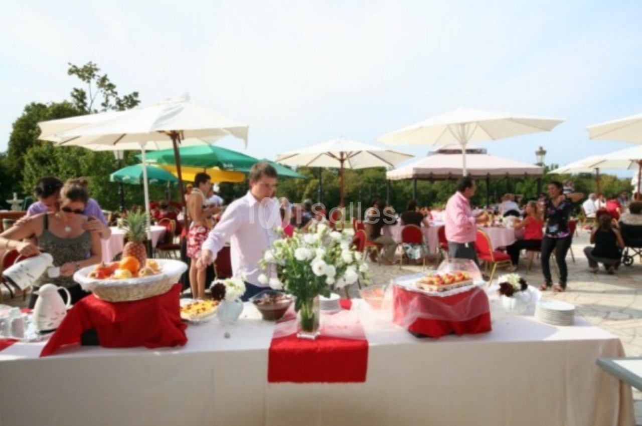 Buffet en plein air avec nappes blanches et rouges, fleurs au centre, invités se servant sous des parasols.