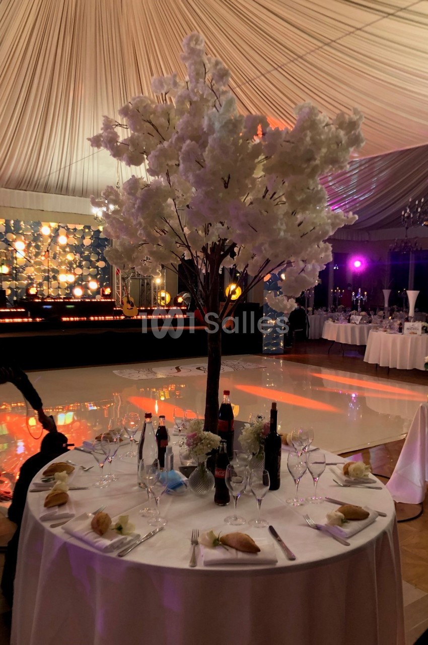 Table dressée avec nappes blanches, couverts, bouteilles et pain, décorée d'un arbre fleuri sous une tente éclairée.