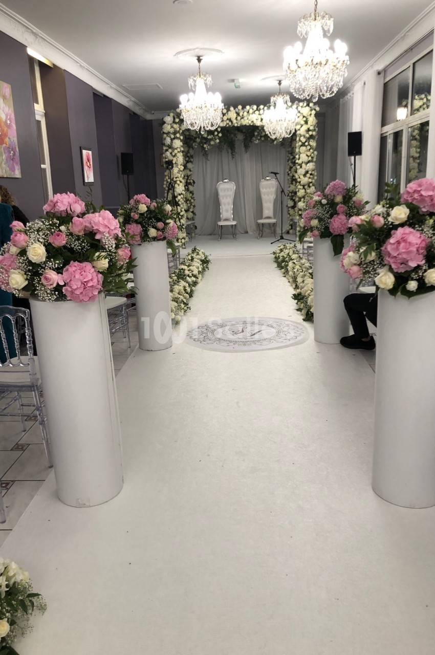 Allée décorée de fleurs blanches et roses menant à une arche florale, dans une salle éclairée par des lustres.