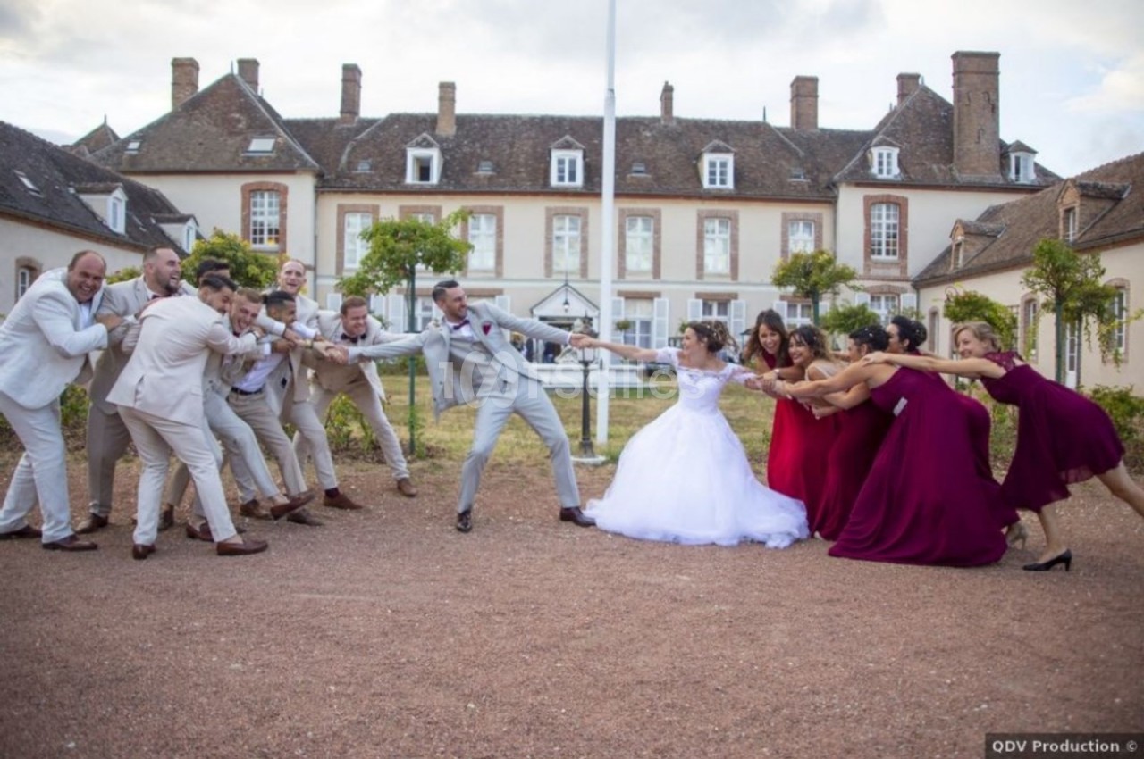 Des invités en tenues de mariage jouent à tirer une corde devant un bâtiment ancien en pierre.