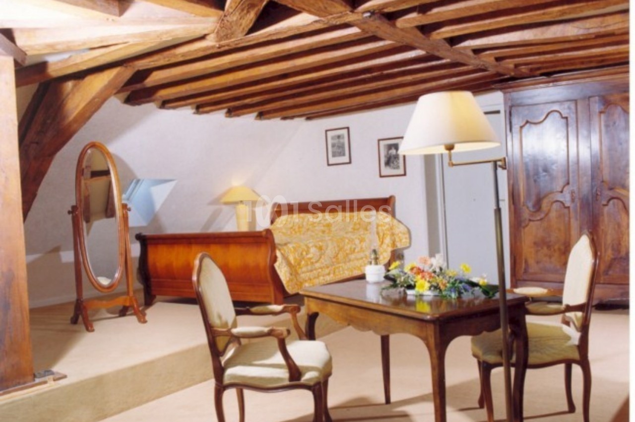Chambre mansardée avec poutres apparentes, mobilier en bois ancien, table, chaises, lit et miroir sur pied.