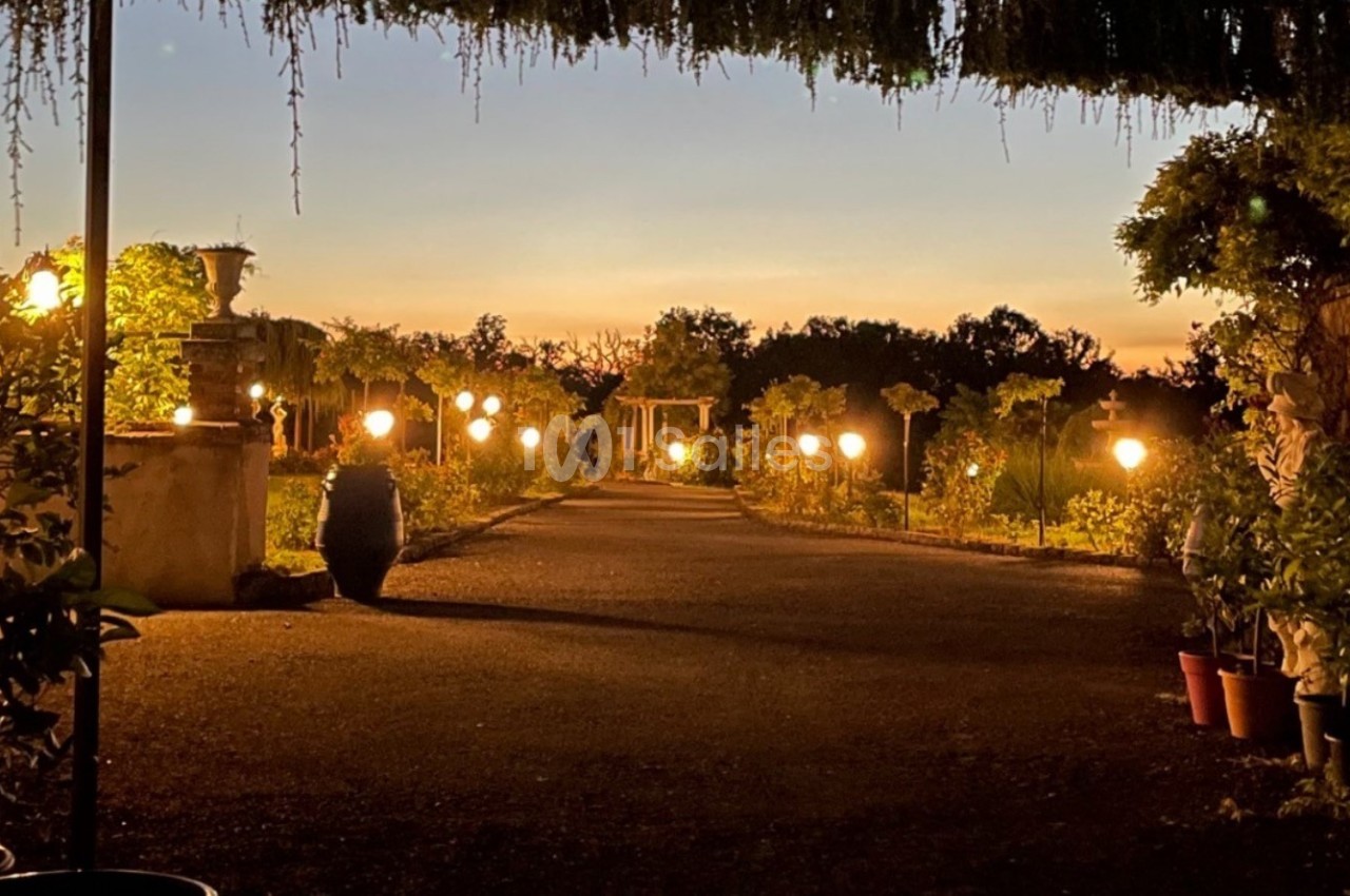 Allée de jardin éclairée par des lampes, bordée de plantes en pot, au crépuscule avec un ciel orangé.