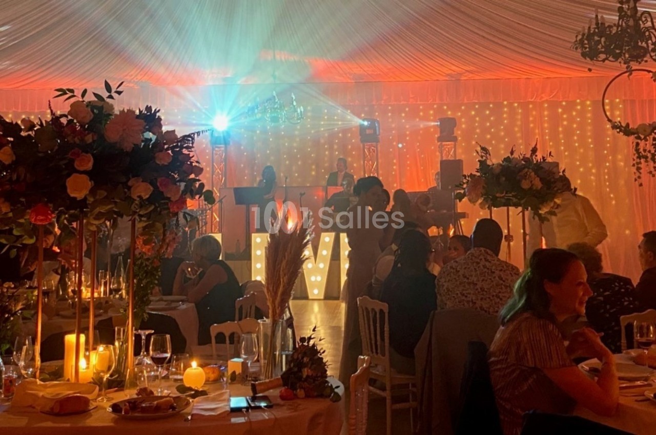 Salle de réception décorée avec des fleurs, éclairage chaleureux et invités assis à des tables lors d'un événement.