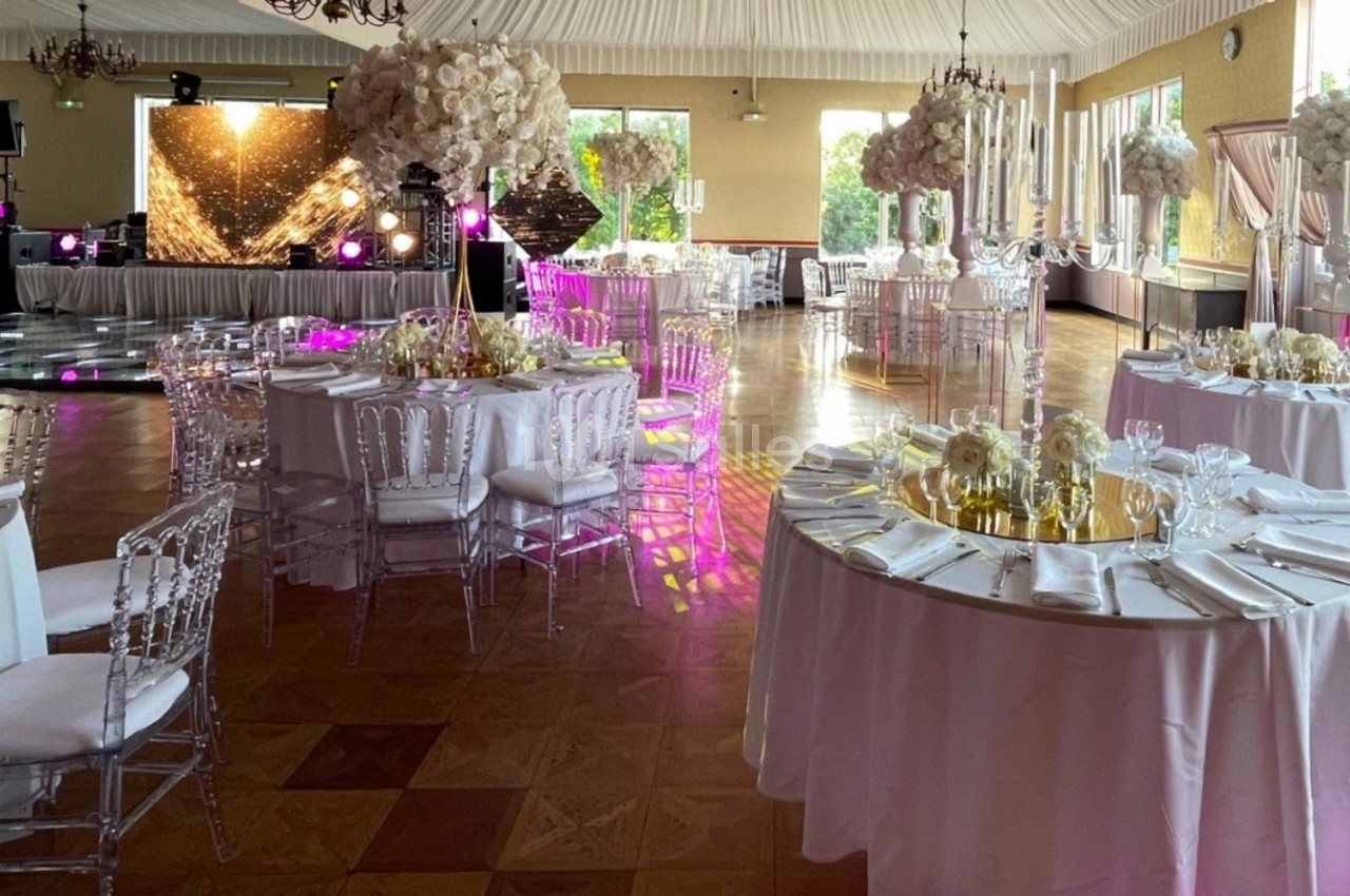 Salle de réception décorée avec des tables rondes, nappes blanches, chaises transparentes et éclairage festif.