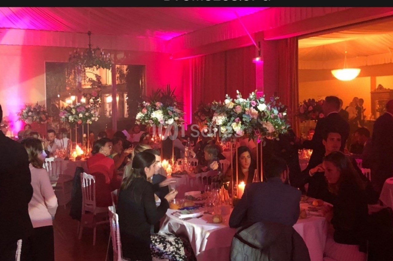 Salle de réception décorée avec des fleurs et éclairée de lumières roses, des invités dînent à des tables rondes.