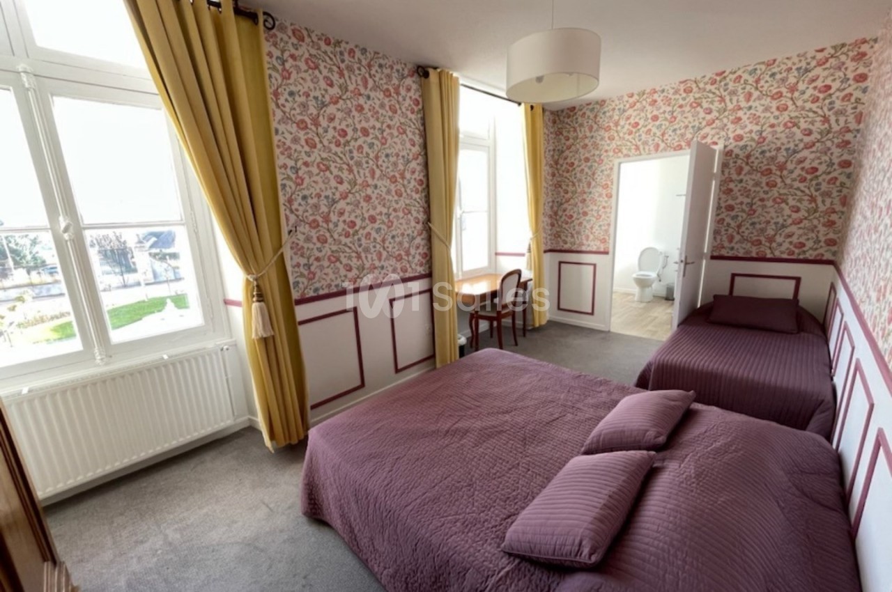 Chambre lumineuse avec deux lits, murs tapissés de motifs floraux, fenêtres avec rideaux jaunes et bureau en bois.