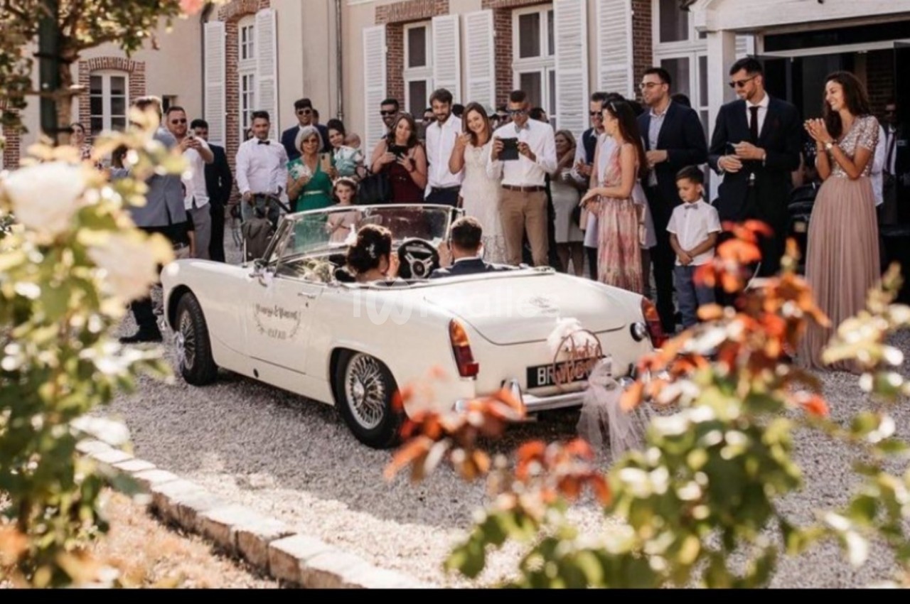 Un couple dans une voiture décapotable blanche quitte une cérémonie entouré d'invités élégants devant un bâtiment.