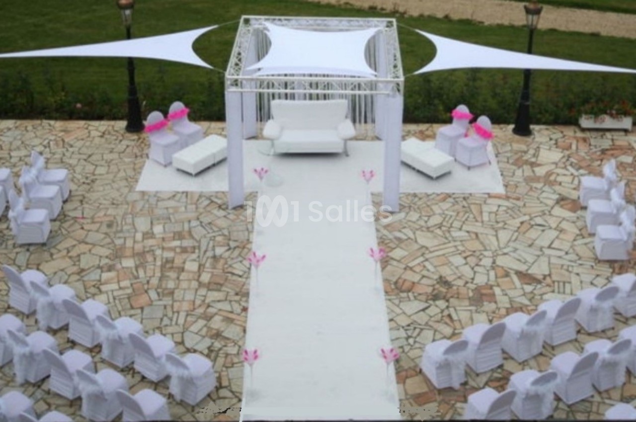 Allée centrale décorée pour une cérémonie en extérieur, entourée de chaises blanches et d'un espace couvert.