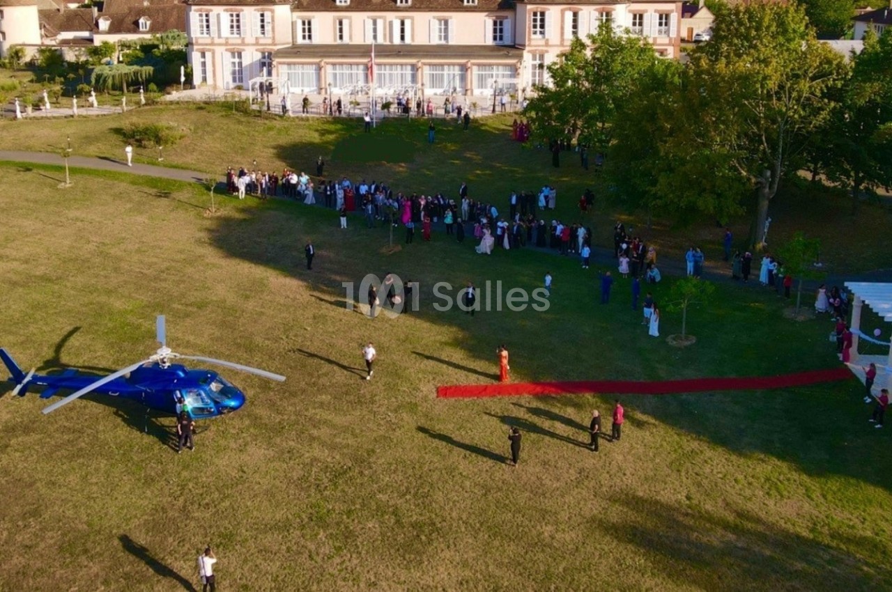 Vue aérienne d'un hélicoptère bleu posé sur une pelouse près d'un bâtiment, avec des invités alignés sur un tapis rouge.