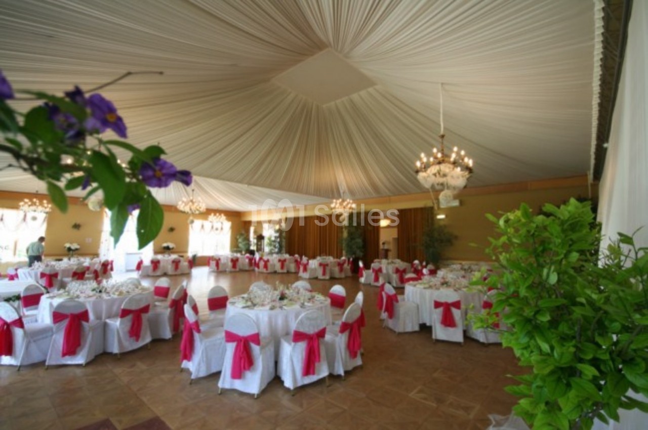 Salle de réception décorée avec des tables rondes, nappes blanches, nœuds roses et lustres suspendus.