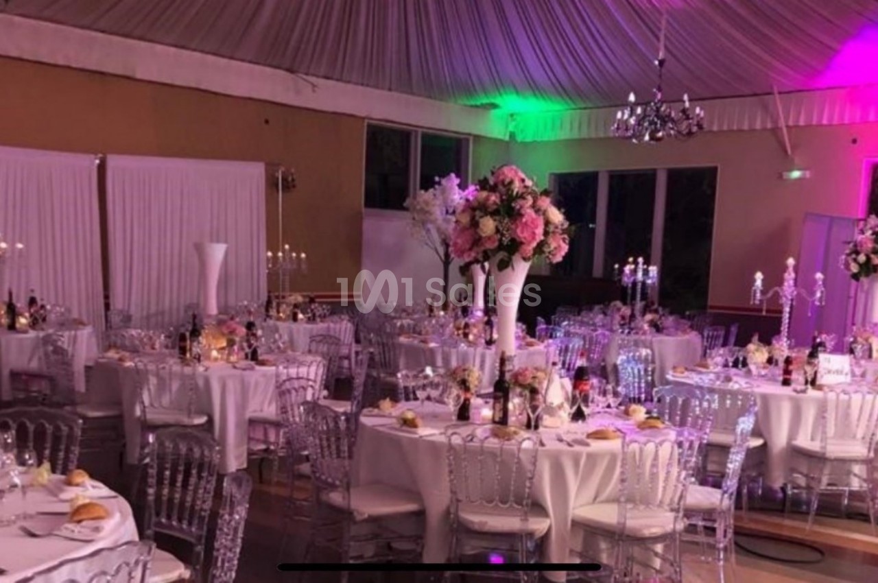 Salle de réception décorée avec des tables rondes, nappes blanches, chaises transparentes et arrangements floraux roses.