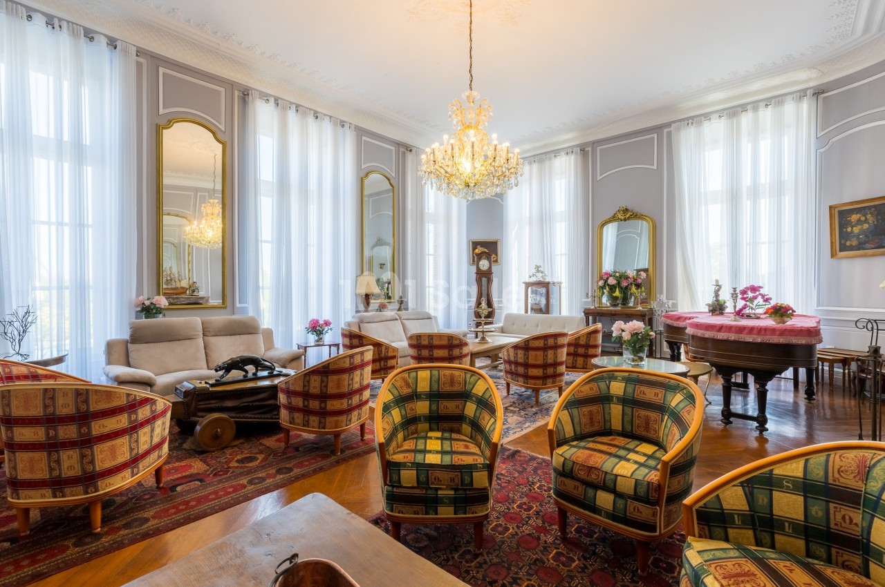 Salon élégant avec fauteuils colorés, lustre central, miroirs dorés, piano et grandes fenêtres aux rideaux blancs.