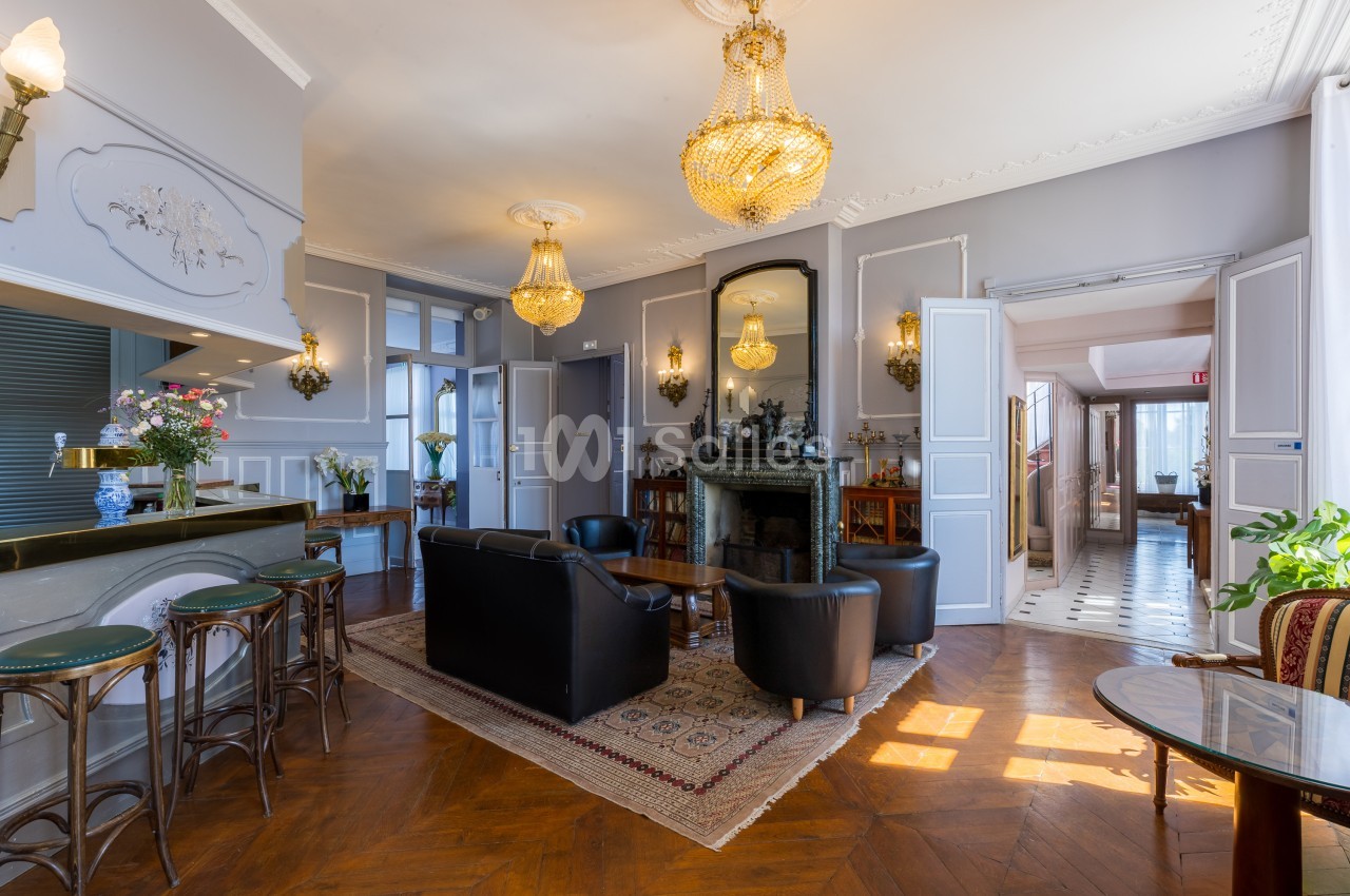 Salon élégant avec parquet, fauteuils en cuir, cheminée, lustres en cristal et lumière naturelle entrant par les fenêtres.