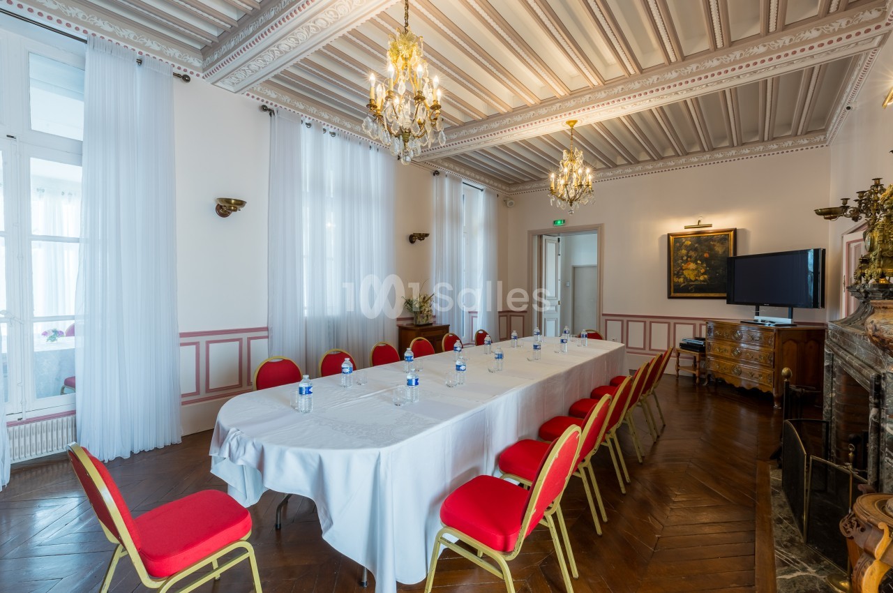 Salle de réunion lumineuse avec table rectangulaire dressée, chaises rouges, lustres et décorations classiques.