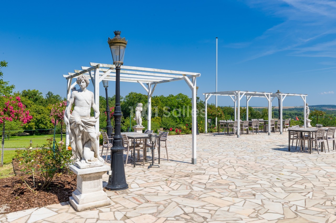 Terrasse pavée avec pergolas blanches, mobilier de jardin, statues classiques et vue sur un paysage verdoyant.