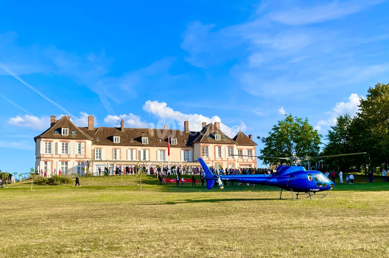 Hélicoptère bleu posé sur une pelouse devant un grand bâtiment historique sous un ciel dégagé.