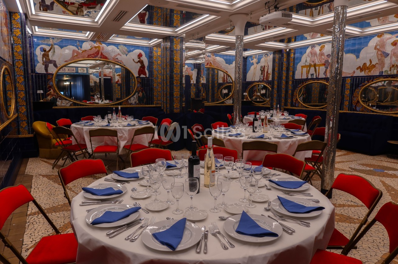 Salle de réception élégante avec tables rondes dressées, nappes blanches, serviettes bleues et miroirs ornés sur les murs.