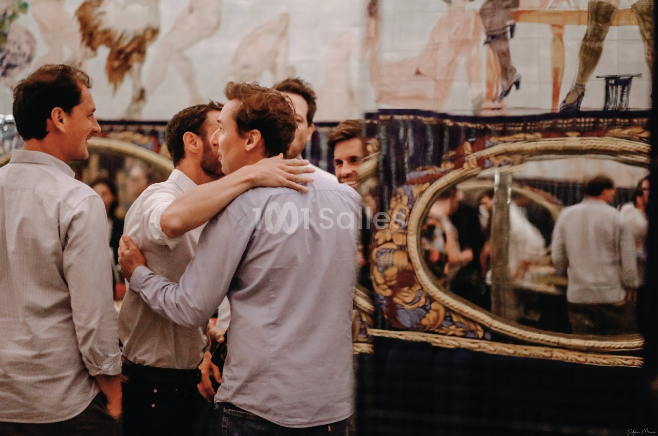 Un groupe d'hommes discutant et s'embrassant dans un intérieur décoré avec des miroirs et des motifs artistiques.