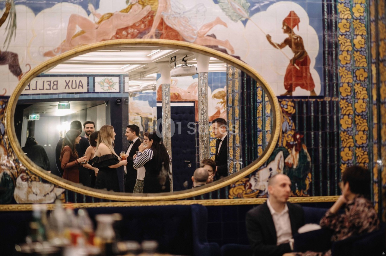 Des personnes discutent dans un espace décoré de fresques colorées, reflétées dans un grand miroir ovale.