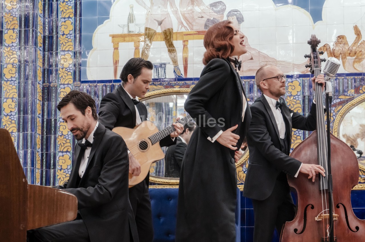 Un groupe de musiciens en tenue élégante joue du piano, de la guitare et de la contrebasse, accompagné d'une chanteuse.
