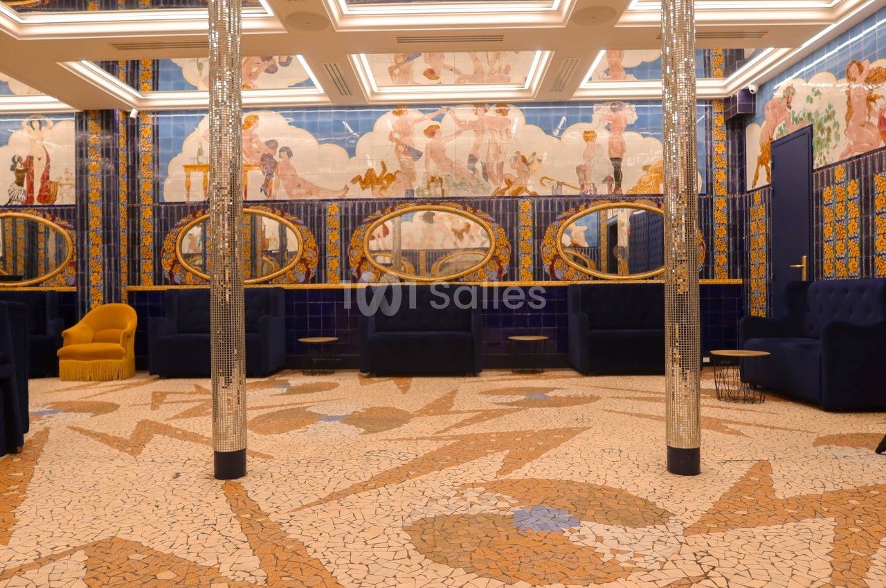 Salle décorée avec des mosaïques au sol, fresques murales colorées, miroirs ovales et fauteuils bleu marine.