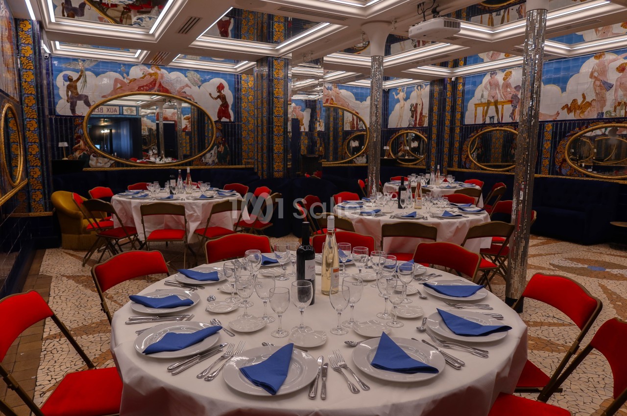 Salle de réception élégante avec tables rondes dressées, chaises rouges et décor mural coloré reflété dans des miroirs…