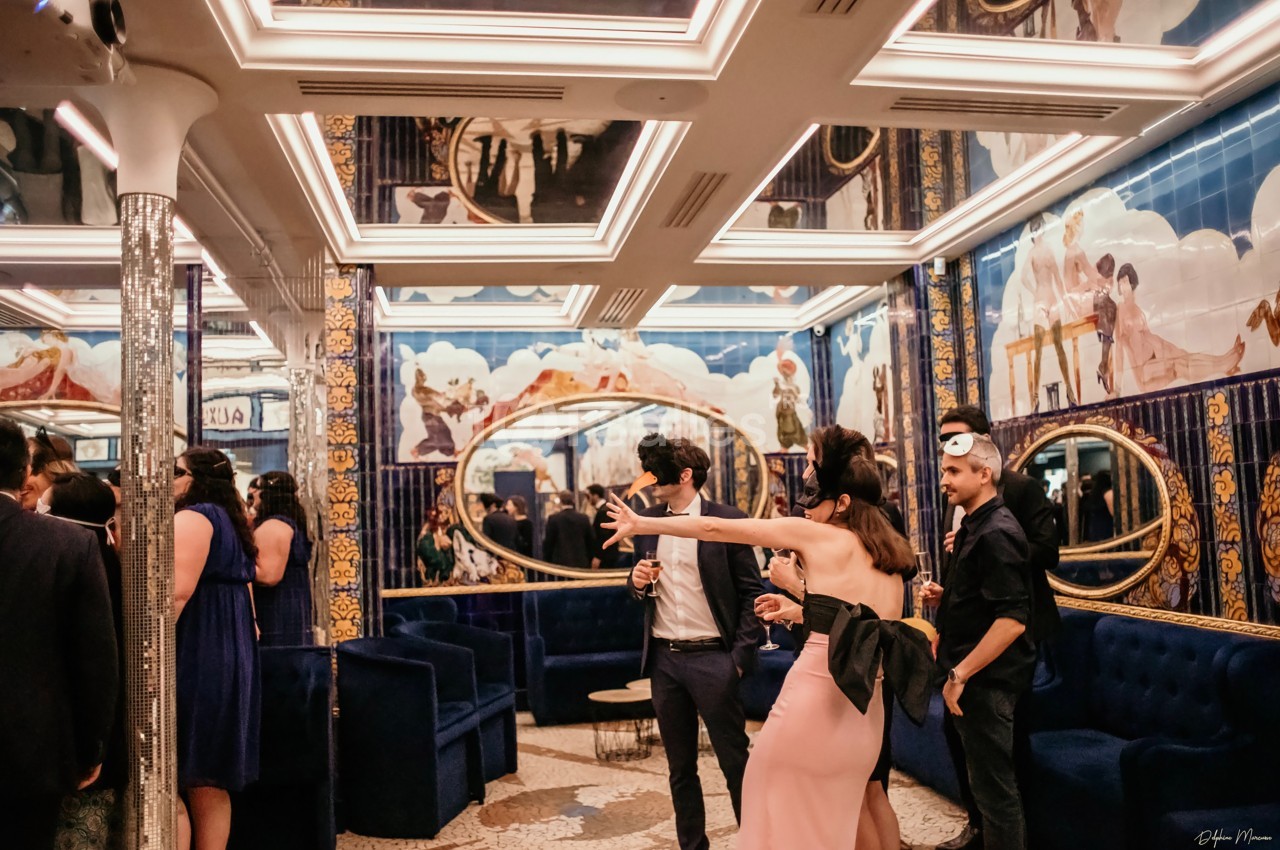 Personnes élégamment vêtues discutant et prenant des photos dans une salle ornée de miroirs et de fresques colorées.