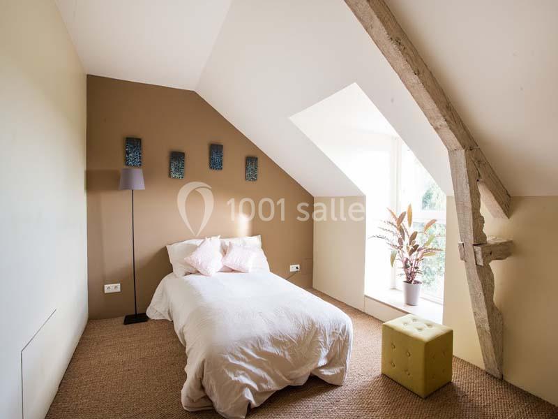 Chambre mansardée avec lit simple, mur beige, poutre apparente, lampe sur pied et fenêtre laissant entrer la lumière.