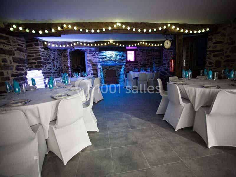 Salle en pierre avec tables rondes dressées, chaises blanches et guirlandes lumineuses suspendues au plafond.