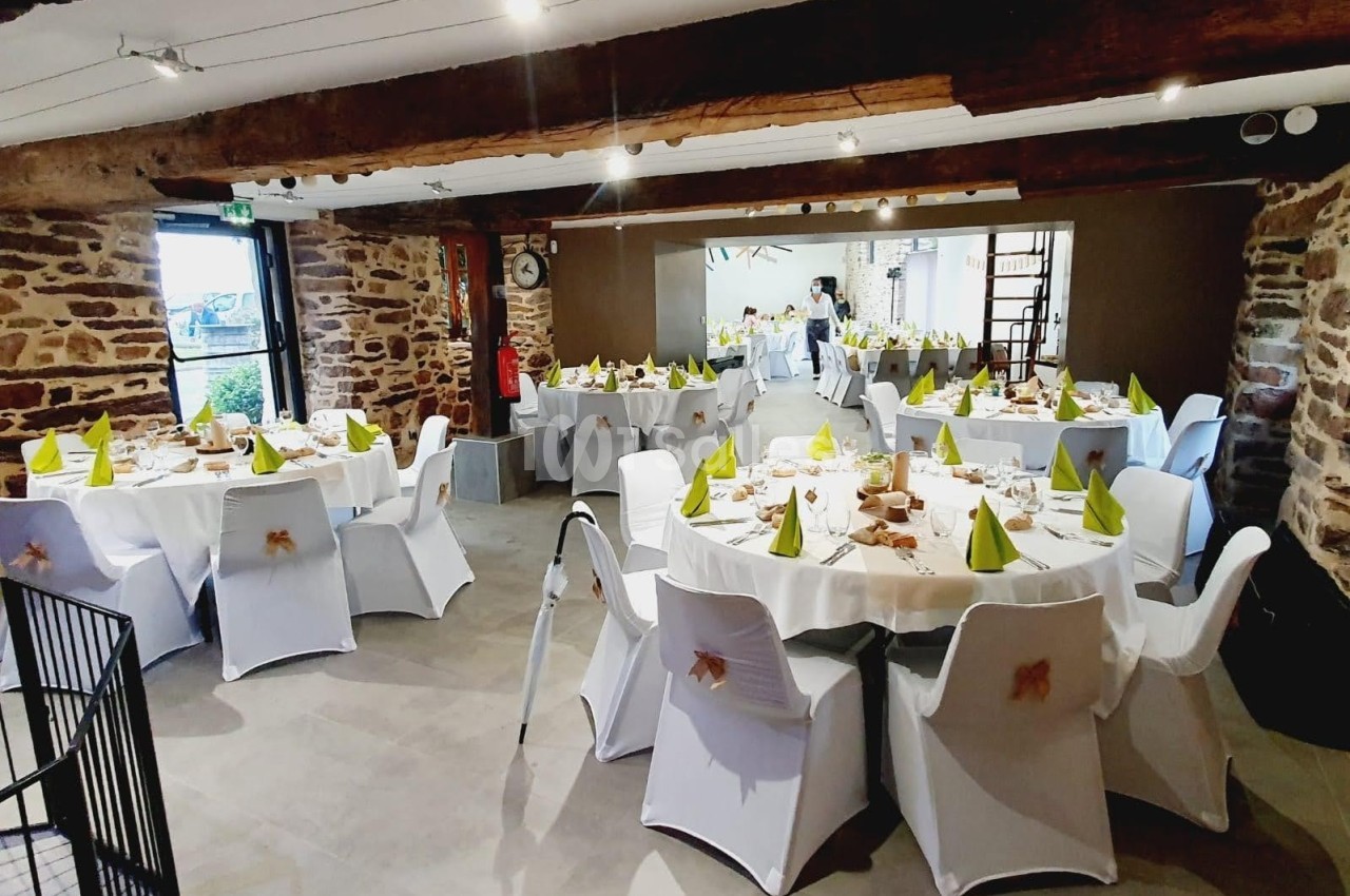 Salle de réception avec des tables rondes décorées de nappes blanches et serviettes vertes, dans un cadre rustique.