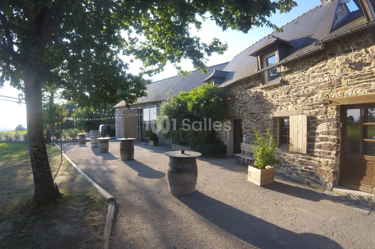 Cour extérieure d'une bâtisse en pierre avec barriques en guise de tables, entourée d'arbres et baignée de lumière.