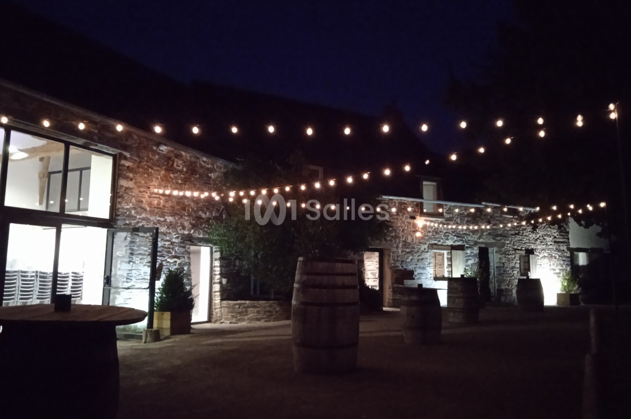 Cour extérieure en soirée avec guirlandes lumineuses suspendues, tonneaux en bois et bâtiments en pierre éclairés.