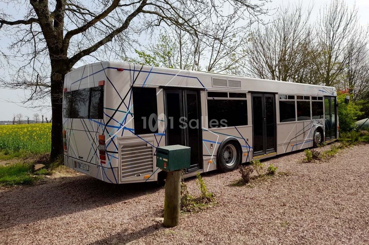 Autobus stationné sur un chemin gravillonné, entouré d'arbres et de champs sous un ciel clair.