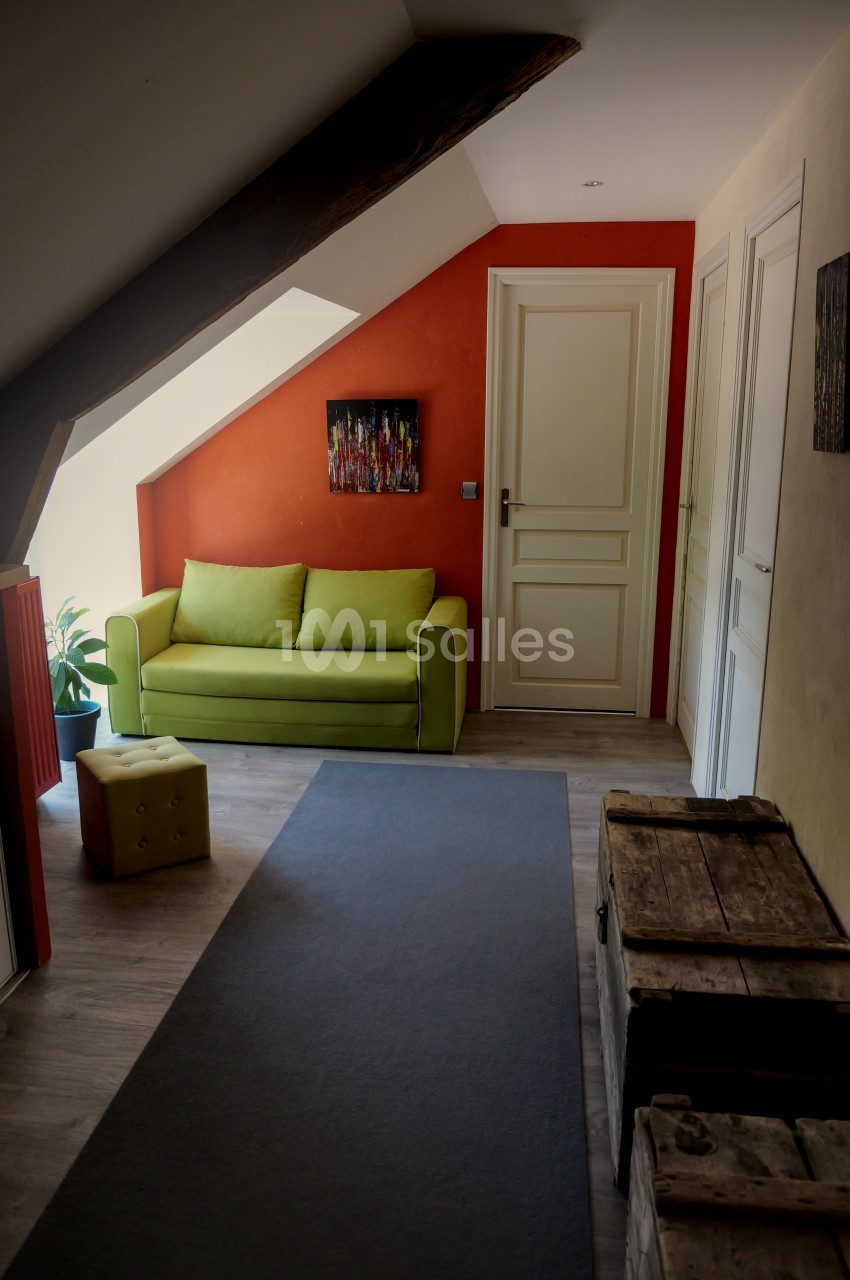 Couloir mansardé avec un mur orange, un canapé vert, des coffres en bois et une lumière naturelle provenant d'une fenêtre.