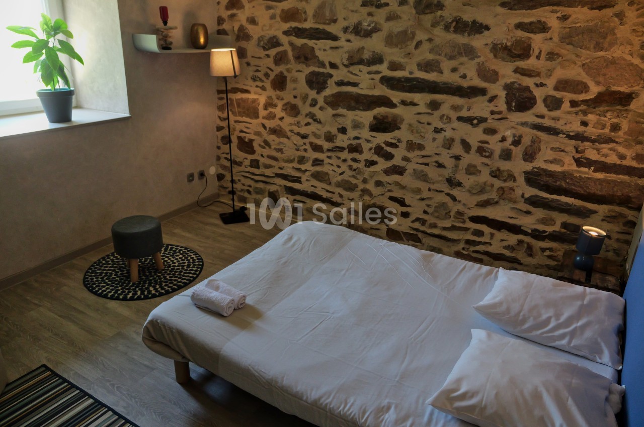 Lit bas avec draps blancs dans une chambre à mur en pierre, éclairée par une fenêtre et une lampe sur pied.