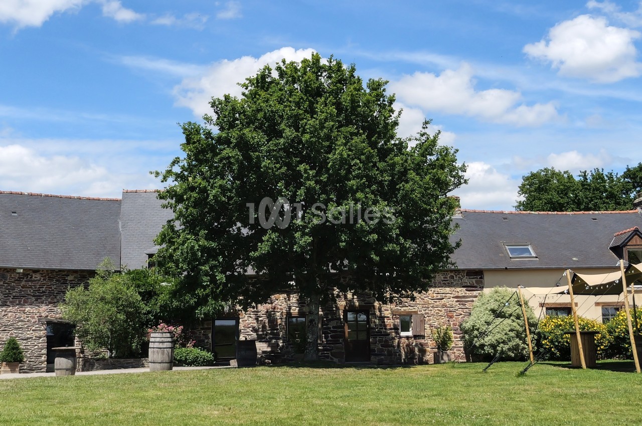 Bâtisse en pierre avec toit en ardoise, grand arbre central et tente beige sur une pelouse sous un ciel bleu.