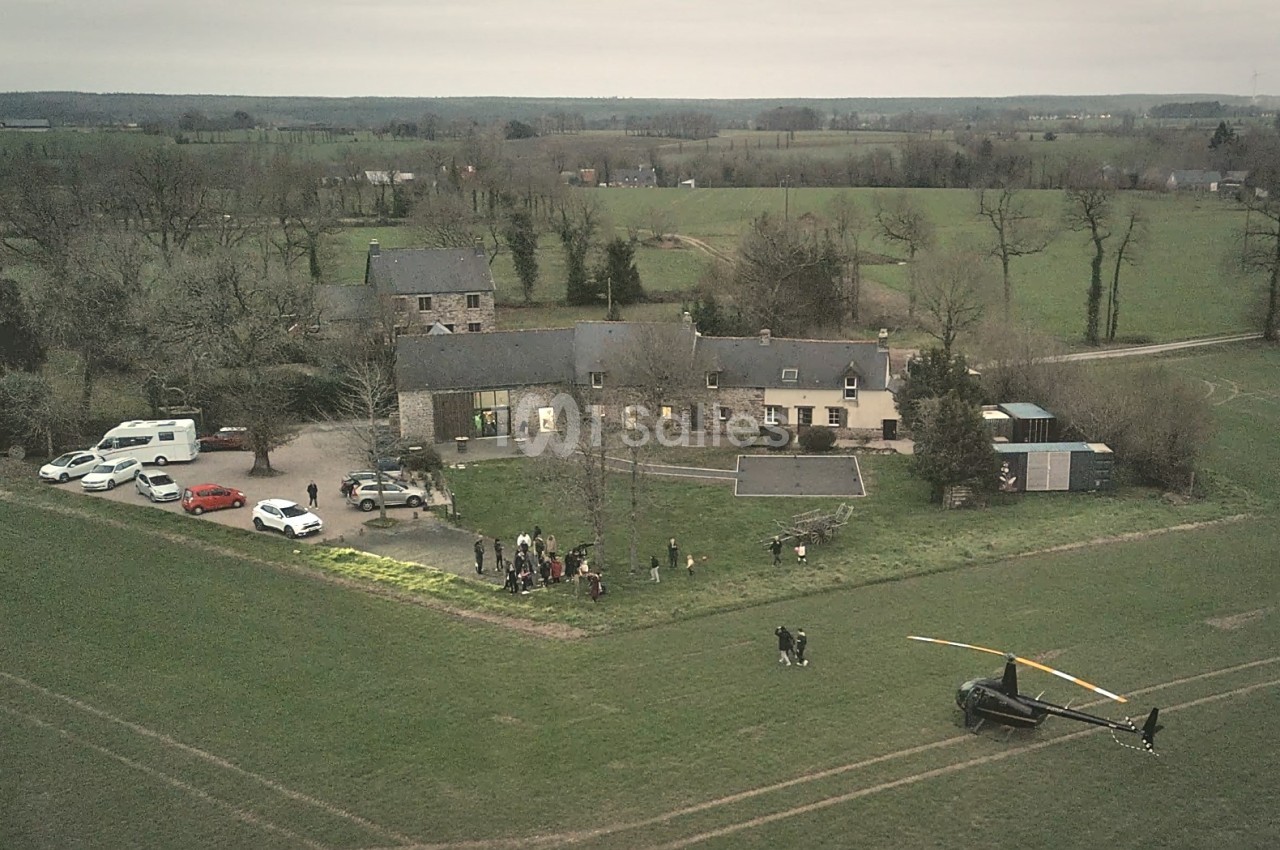 Vue aérienne d'une propriété rurale avec plusieurs bâtiments, des voitures garées et un hélicoptère posé sur un champ.