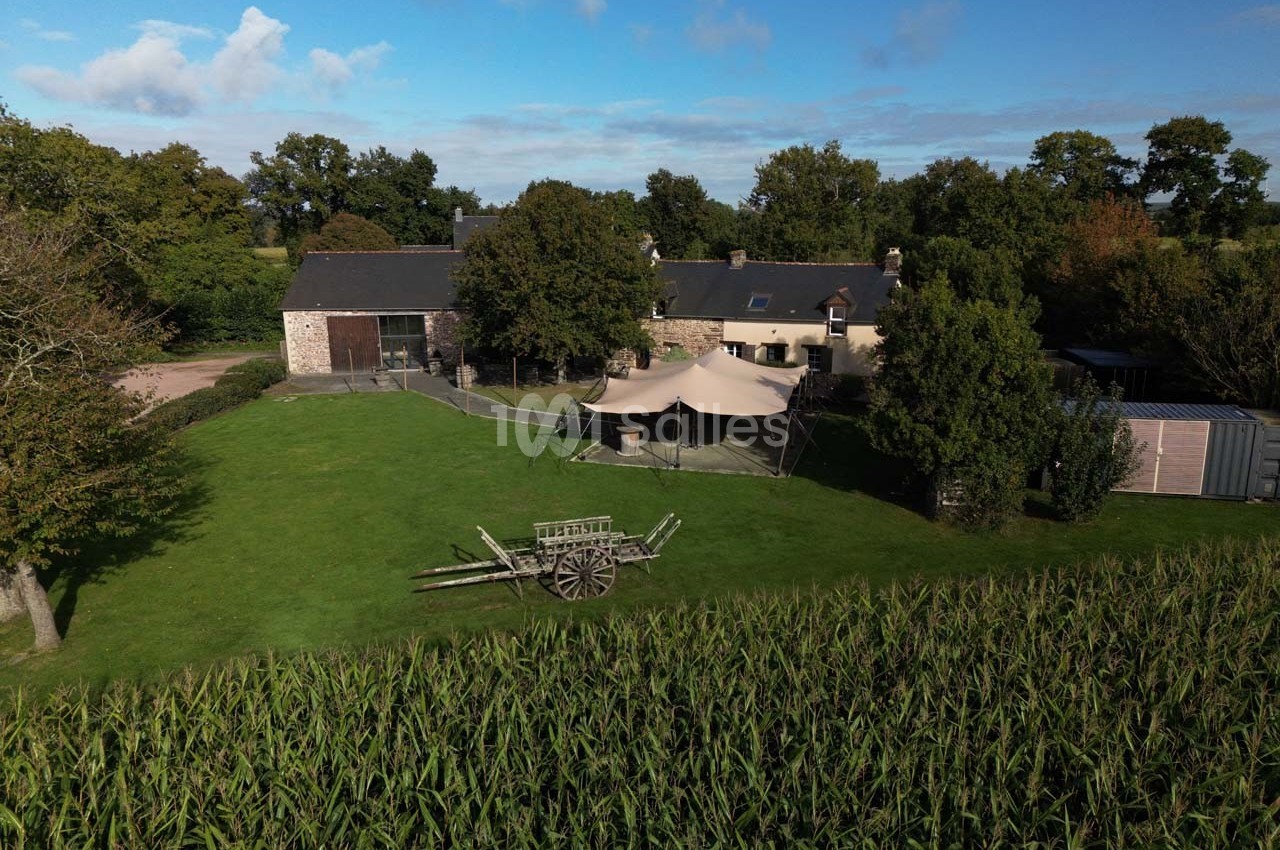 Vue aérienne d'une ferme en pierre avec une tente sur une pelouse, entourée d'arbres et d'un champ de maïs.