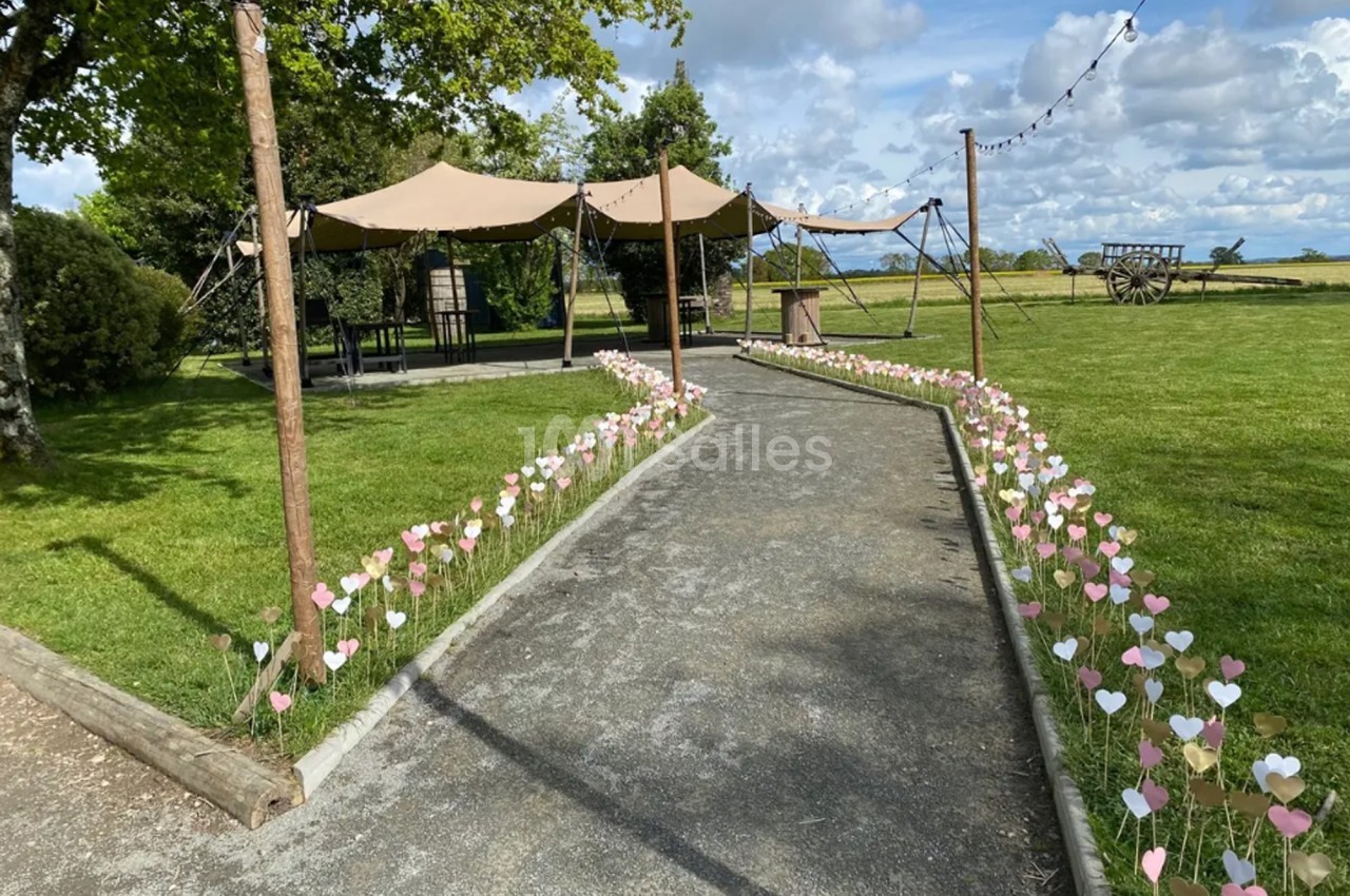 Allée bordée de petits cœurs en papier menant à des tentes beige dans un espace vert sous un ciel partiellement nuageux.