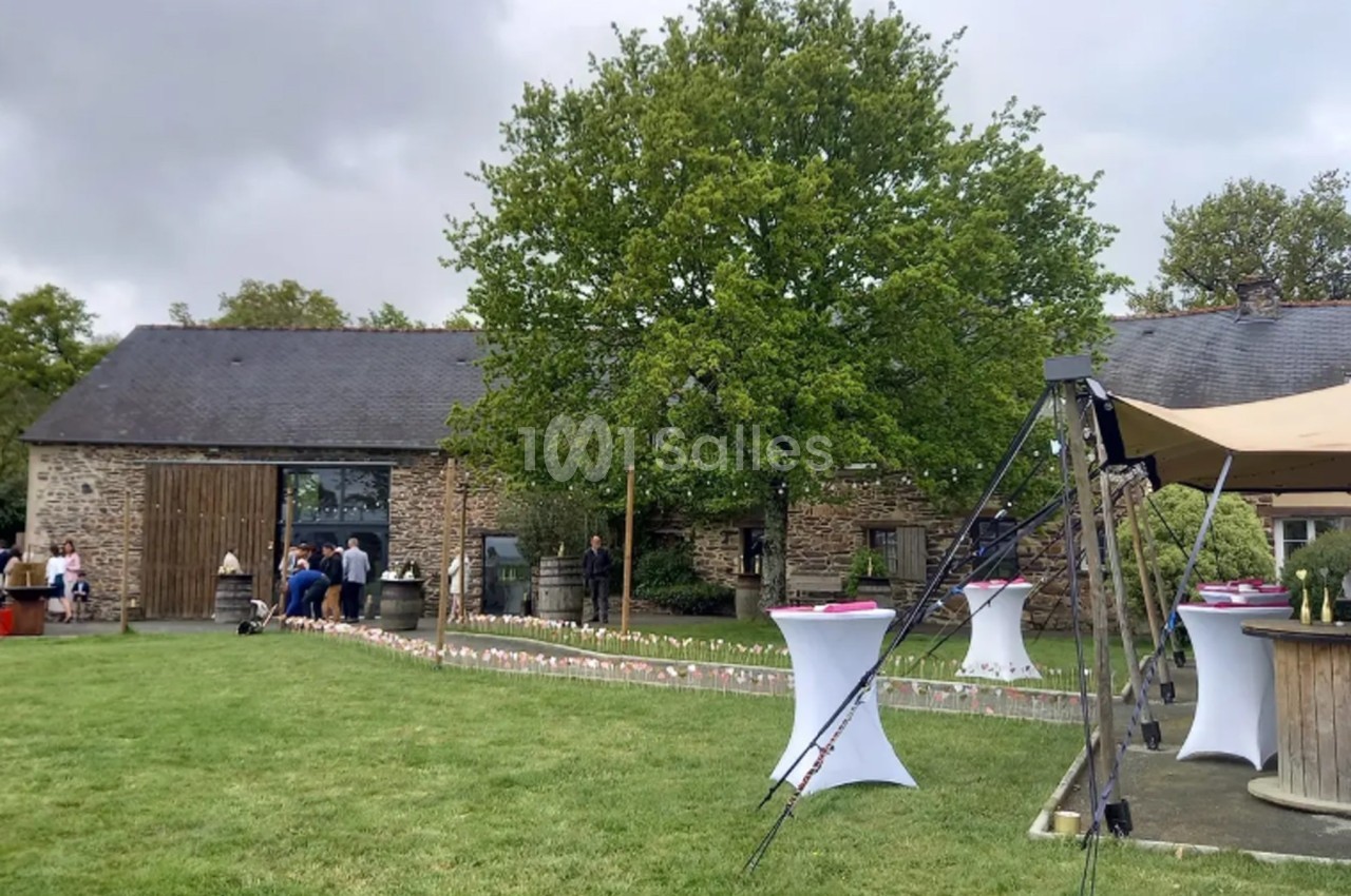 Extérieur d'une bâtisse en pierre avec pelouse, tables hautes blanches et décorations pour un événement en plein air.