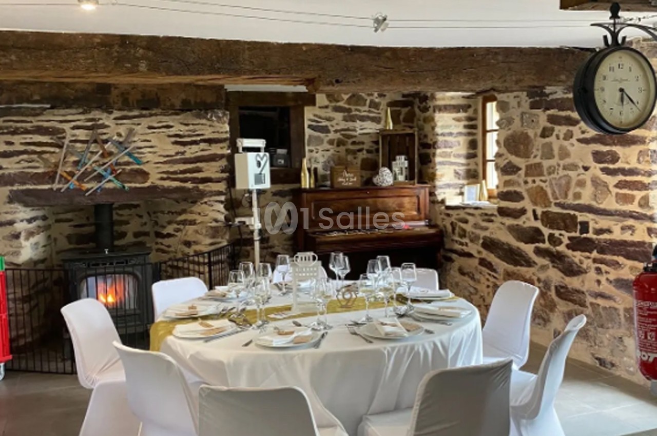 Salle rustique avec murs en pierre, table ronde dressée pour un repas, poêle allumé et piano en arrière-plan.