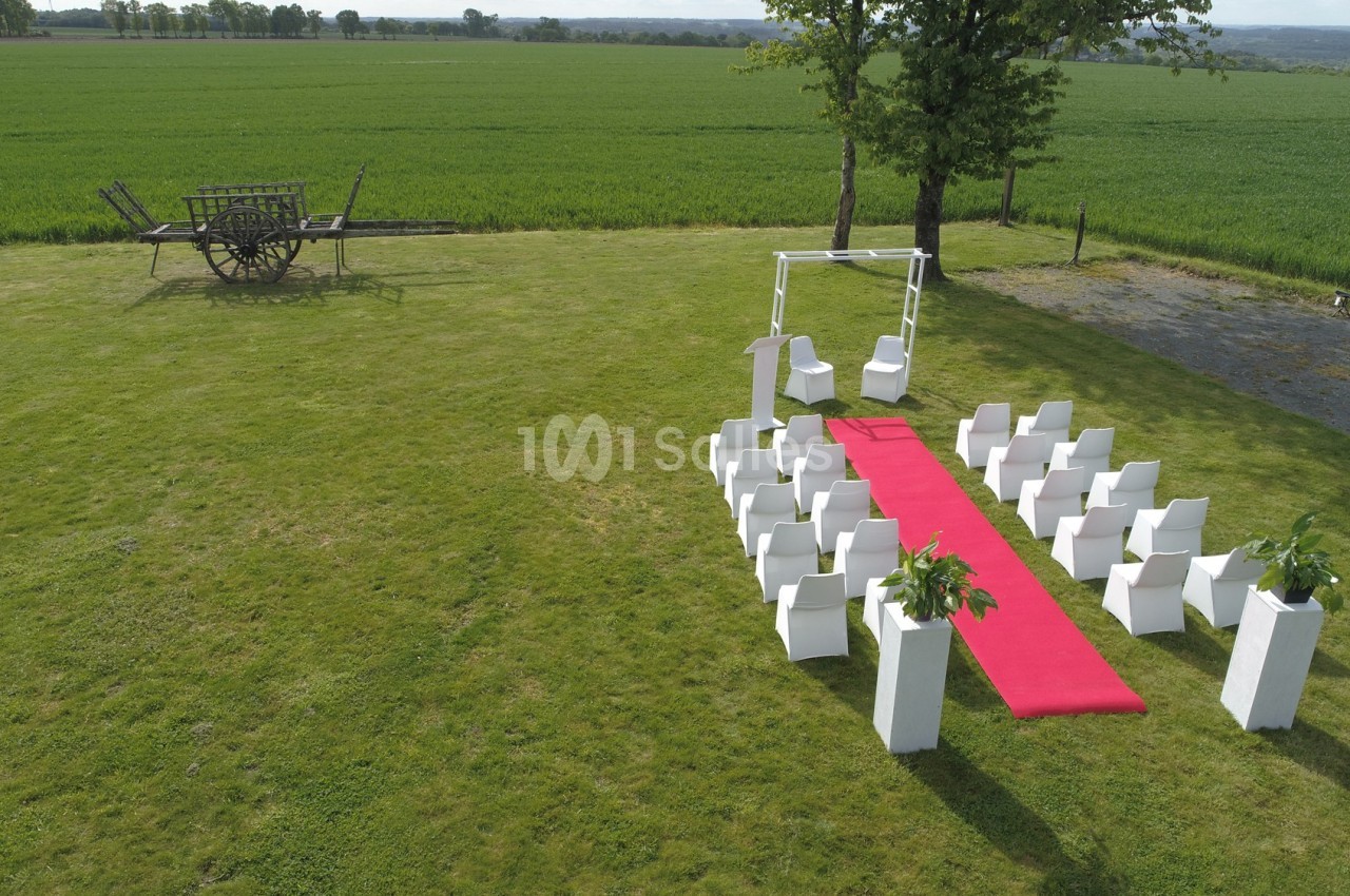 Chaises blanches disposées en extérieur autour d'une allée rouge, avec un arche décoratif et un champ en arrière-plan.