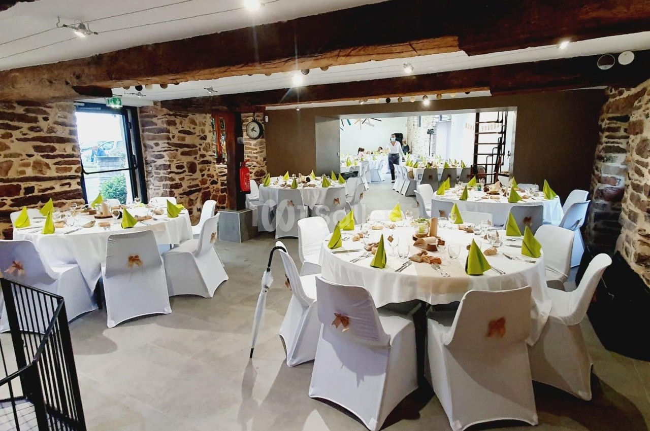 Salle de réception décorée avec des tables rondes, nappes blanches, serviettes vertes et murs en pierre apparente.